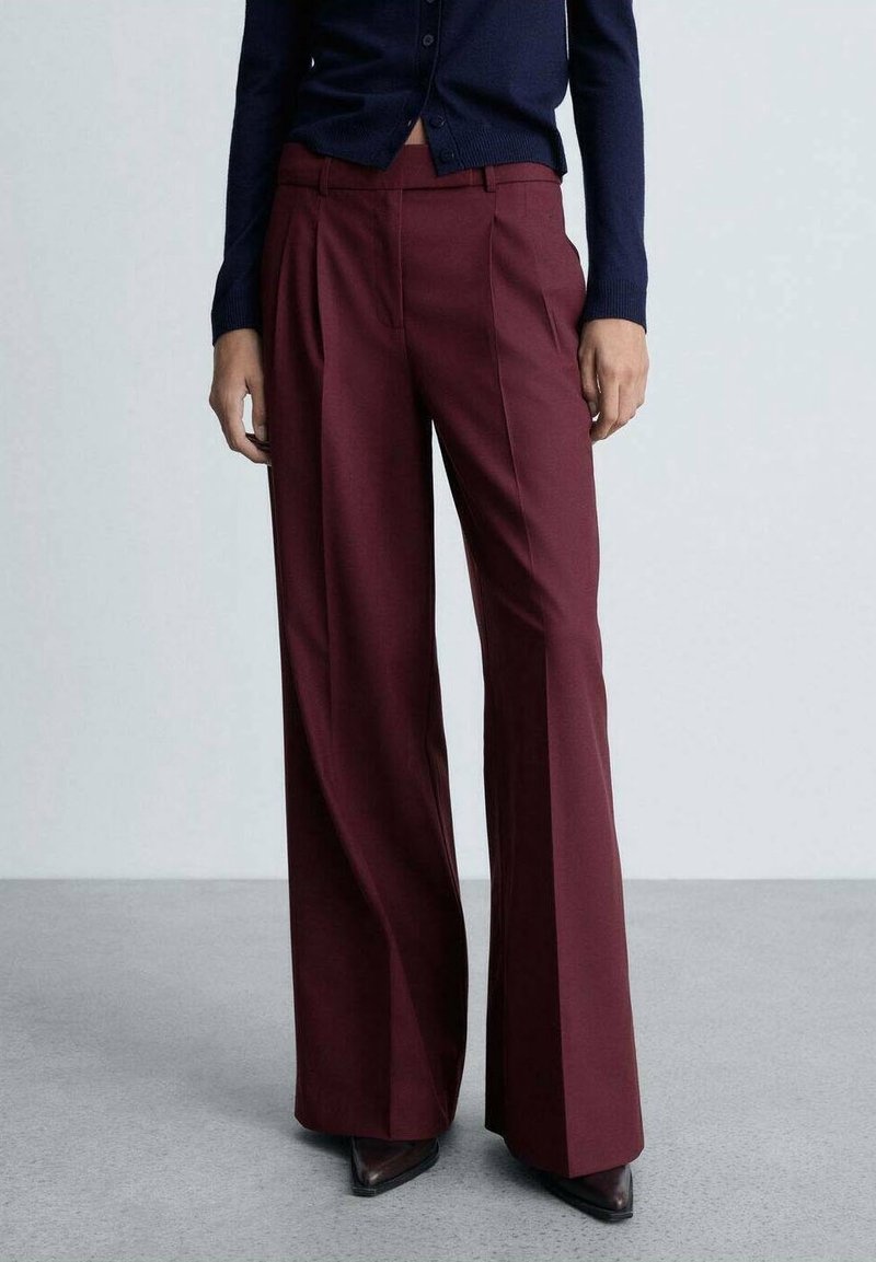 Personne portant un pantalon bordeaux taille haute à jambes larges, associé à un pull bleu marine et des chaussures pointues foncées, debout sur un sol gris.