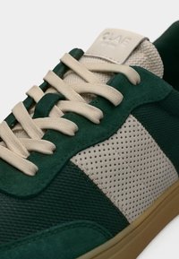 Πράσινο και μπεζ αθλητικό παπούτσι με κρέμ κορδόνια, πλευρική πάνελ με τρύπες, υφασμάτινες και σουέτ υφές, και λεπτομέρεια στη σόλα γόμας.
