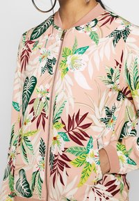 Chaqueta bomber floral en rosa claro con patrones de hojas en verde y burdeos. Presenta un cierre con cremallera y puños de canalé. Textura de tela suave.