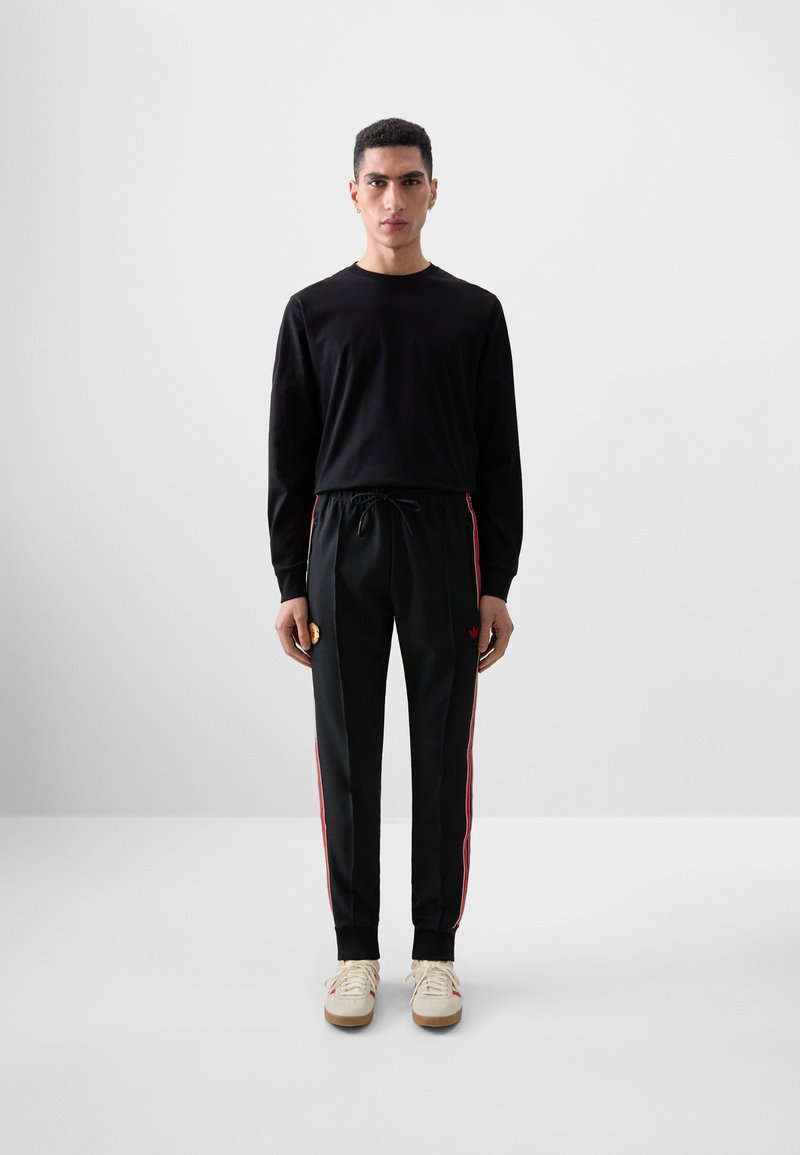 Chemise noire à manches longues associée à un pantalon noir avec des bandes latérales rouges. Modèle portant des chaussures beiges se tient devant un fond gris uni.