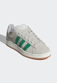 Szare zamszowe sneakersy z trzema zielonymi paskami, okrągłym noskiem, płaską podeszwą, sznurowanym przodem oraz białym logo na języku. Teksturowana cholewka i gumowa podeszwa.