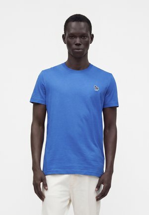 Mand iført en blå t-shirt med korte ærmer med et lille zebra-logo på brystet og lyse bukser, stående mod en ensfarvet baggrund.