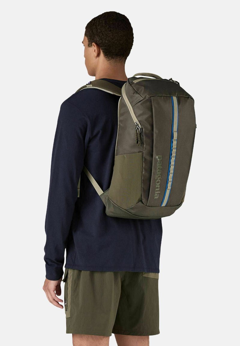 Zaino verde con striscia blu, realizzato in tessuto durevole. Presenta spallacci imbottiti e logo sul lato. Vano con cerniera visibile.