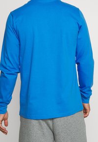 Mies, który ma na sobie jasnoniebieską koszulę z długim rękawem i jasnoszare spodnie, pokazany od tyłu na jednolitym tle.