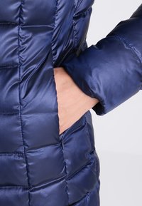 Doudoune bleu marine à motif matelassé, tissu brillant et poche latérale zippée, avec une main visible dans la poche.