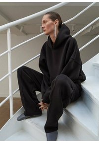 Zwarte oversized hoodie en bijpassende sweatpants met een zachte textuur. Opvallende lange rechthoekige oorbellen. De scène bevat een witte trap.