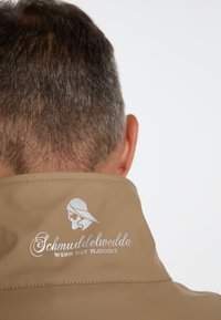 Ljusbrun vattentät jacka med hög krage, slät textur och silvertryckt logotypdetalj på ryggen med karaktärsdesign.