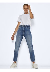 Hochtaillierte, schmale blaue Jeans aus Denim mit fünf Taschen und traditioneller Steppnaht, kombiniert mit einem weißen Kurzarmhemd und Sneakern.