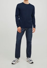 Mężczyzna ubrany w granatowy sweter z długim rękawem, ciemnoniebieskie dżinsy oraz białe, czarne i szare sneakersy, stojący na tle jednolitego jasnego tła.