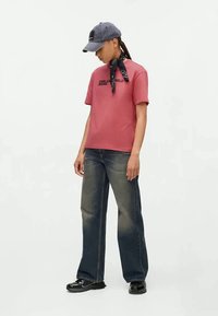 Pink cotton t-shirt with short sleeves, black logo print. Dark blue denim wide-leg jeans and a gray cap with a contrasting bandana tied around the neck.