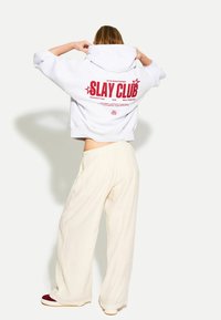 Svetle šedá oversize mikina s výrazným červeným textem "SLAY CLUB" na zádech, v kombinaci s volnými krémovými kalhotami.