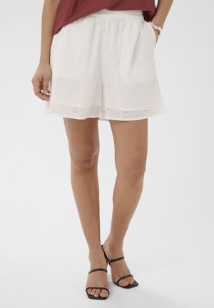 Kvinde iført hvide shorts med hulmønster, en vinrød top, sorte sandaler med åbne tæer og stropper samt et sølvarmbånd på venstre håndled.