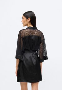 Mustast satiiniesiosaga ning läbipaistva geomeetrilise pitsiga varrukatega rüü. Varustatud lipsuga vöökohal ja põlveni ulatuva lõikega.