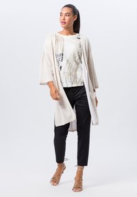 Cardigan en maille beige clair avec des manches larges, portée sur un top à motifs noir et blanc, associée à un pantalon noir fuselé et des sandales à lanières.