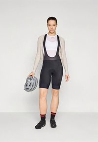 Černé cyklistické šortky s vycpávkou na sedadle, bílá přiléhavá košile, béžový svetr, černé boty a šedá helma držená v ruce.