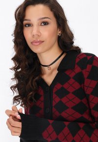 Čierny a červený cardigan s argyle vzorom, s golierom a piatimi gombíkmi; má rebrované manžety a mäkkú pletenú textúru.