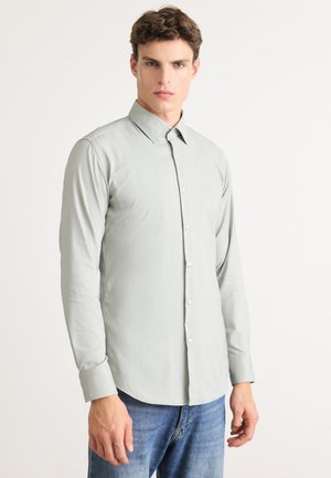 Junger Mann, der ein hellgraues Langarmhemd mit Knöpfen und blaue Jeans trägt, steht vor einem schlichten weißen Hintergrund.