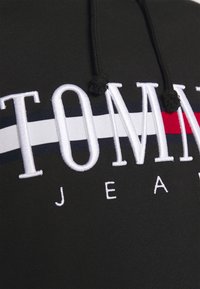 Sudadera negra con capucha, que presenta un logo en letras bordadas "TOMMY JEANS" en colores blanco, rojo y azul marino. Detalle de cordón en la capucha.