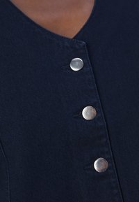 Ναυτικό τζιν ένδυμα με V-λαιμόκοψη και πέντε μεταλλικά κουμπιά. Μαλακή υφή με εμφανή ραφή και ελαφρώς δομημένο σχήμα.