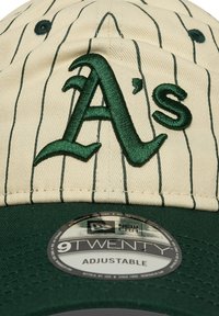 Gorra crema con rayas verdes, que presenta un logo "A's" bordado en verde elevado. Ajuste ajustable con un visera plana verde. Marca New Era visible.