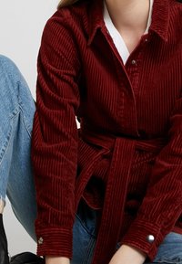 Rode corduroy shirt met een gestrikt riempje, voorzien van een knoopsluiting op de kraag en een ribbelstructuur. Gekoombineerd met blauwe jeans. Zwarte laarzen zichtbaar.