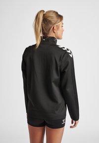 Zwarte zip-up jas met hoge kraag, met witte chevronstrepen op de schouders. Gladde structuur en aansluitend ontwerp. Casual atletische stijl.