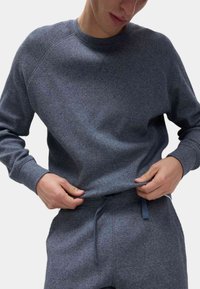 Szary sweter z dzianiny o teksturowanej powierzchni i ściągaczach na rękawach. Posiada okrągły dekolt i luźny krój, zestawiony z pasującymi spodniami.