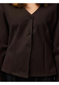 Smeđa bluza s gumbima i V-izrezom, ukrašena s tri tamna gumba i prilagođenim krojem. Glatki materijal, presavijena rukava i rub koji završava u visini struka.