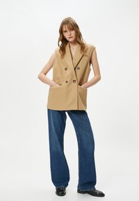 Blazer senza maniche beige con design doppiopetto e tasche frontali, abbinato a pantaloni in denim blu larghi e scarpe nere.