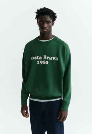 Mladý muž s tmavou pokožkou a kučeravými vlasmi, ktorý nosí zelenú mikinu s bielym textom "Costa Brava 1980" a tmavomodré džínsy.