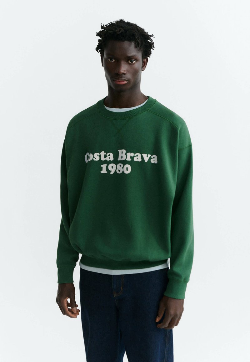 Jeune homme à la peau foncée et aux cheveux bouclés portant un sweat-shirt vert avec un texte blanc "Costa Brava 1980" et un jean bleu foncé.