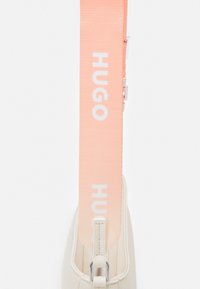 Beige faux leather bag with a smooth texture, featuring a wide pink strap embossed with "HUGO" in white. Zipper closure at the top.