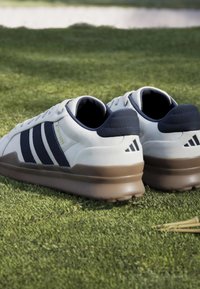 Λευκά και ναυτικά sneakers με λαστιχένιες σόλες στο γρασίδι, που δείχνουν τις ρίγες της Adidas και το λογότυπο στη φτέρνα, από γωνία πίσω αριστερά.