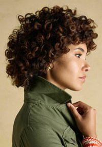 Žmogus su garbanotais plaukais laiko alyvuogių žalios striukės apykaklę, dėvi žvaigždės formos auskarą ir raudonai baltą austą apyrankę.