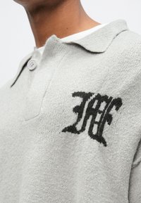 Ανοιχτό γκρι μακρυμάνικο πόλο από υφασμάτινη ύφανση, με κουμπωτό κλείσιμο και μαύρο κεντητό λογότυπο στο στήθος.