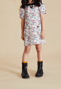 Robe fleurie avec de courtes manches bouffantes, base blanche, fleurs roses et bleues, col noir et bottes noires accentuées de jaune.