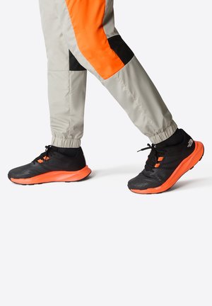 Baskets de course noires et sportives avec des accents oranges et une tige noire texturée. Semelle légère avec un midsole orange audacieux. Pantalons gris avec un panneau orange.