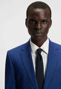 Blauw pak met een notch lapel, pasvorm silhouet, gecombineerd met een wit overhemd met blauwe bloemenpatronen en een effen zwarte stropdas.