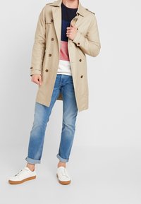 Beige trenchcoat met dubbelrijige knopen en schouderepauletten, gecombineerd met een kleurgeblokte trui, blauwe jeans en witte sneakers.