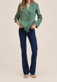 Grün gemusterte Bluse mit V-Ausschnitt und Knopfverschluss, kombiniert mit blauen, ausgestellten Jeans. Modell trägt weiße Absatzsandalen. Schlichter Hintergrund.