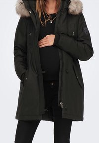 Svart parka-jacka med pälsbeklädd huva, knäppning framtill och sidofickor, båda bärs över svarta kläder, med ett märke på ärmen.