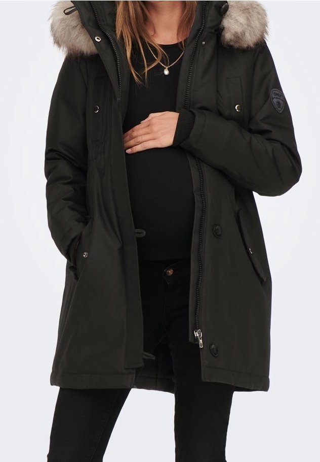 Svart parka-jacka med pälsbeklädd huva, knäppning framtill och sidofickor, båda bärs över svarta kläder, med ett märke på ärmen.