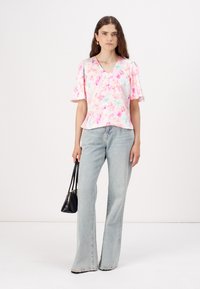 Blusa floral en rosa y blanco con mangas cortas y escote en V. Combinada con jeans acampanados en azul claro y sosteniendo un pequeño bolso negro.