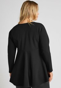 Musta pitkähihainen paita, joka on valmistettu tekstuurisesta materiaalista. Paita on vartalonmyötäinen ja siinä on levenevä helma. Pyöreä pääntie ja yksinkertainen muotoilu.