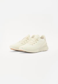 Lys beige sneakers med en teksturert strikkoverdel, har flate lisser, rund tå og en gummisåle med lett mønster.