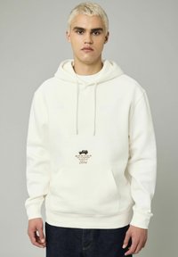 Un hoodie à col rond en tissu crème, doté d'une poche kangourou à l'avant, de cordons de serrage et d'un logo Ford Bronco avec un accent textuel.