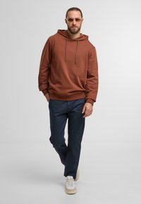 Brauner Kapuzenpullover mit Kordelzug, kombiniert mit dunkelblauen Hosen und hellen Turnschuhen. Weicher Stoff und lässiger Schnitt schaffen einen entspannten Look.