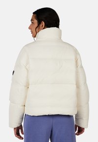 Giacca a vento bianca con colletto alto, caratterizzata da sezioni trapuntate, polsini elasticizzati e una vestibilità rilassata, abbinata a pantaloni sportivi blu.