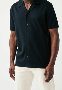 Camicia nera a maniche corte con bottoni, realizzata in un tessuto testurizzato con strisce verticali. Progettata con colletto e bottoni neri, indossata con pantaloni chiari.
