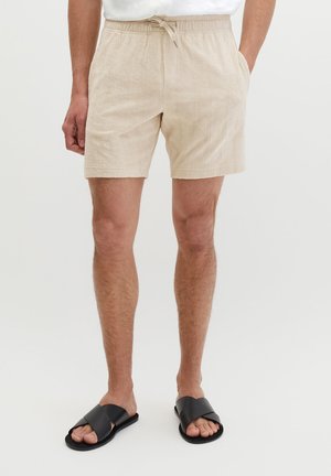 Mężczyzna noszący beżowe, teksturowane szorty ze ściągaczem, kieszeniami bocznymi oraz czarne klapki na stopie z krzyżującymi się paskami na jednolitym tle.